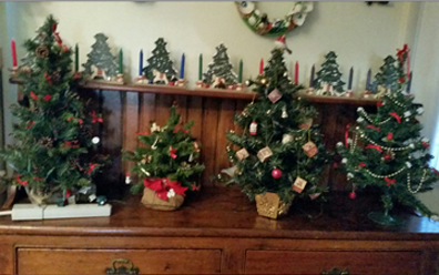 On a sideboard are three conifers with xmas decorations that are about 14 inches tall, Also shown is another that is about 7 inches tall. They are artificial Christmas trees in miniature.  Behind and above on a shelf are seven little 6-7 inch high coniferous trees, each with tiny decorations and flanked by 4 inch candles. 