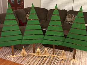Four green-painted wooden slats on a vertical brown sturdy stick supported to stand as unadorned Christmas trees on stands, standing about 4.5 feet tall, plus 4 small trees, same as large, but about 8 inches tall, attheir feet.