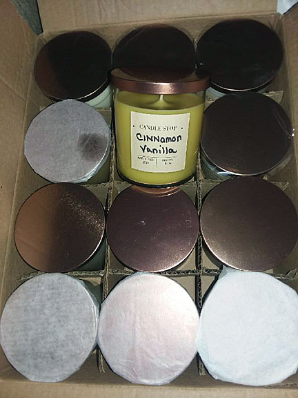 A cardboard box has 12 jars of candles, each with a lid. One jar is pulled out so we can read that it says "Cinnamon".