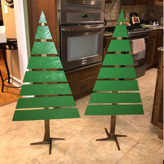 Two unadorned wooden "Christmas trees", made out of green-painted horizontal slats of 2 by 4s nailed to a brown-painted vertical 3 x 3 stick.  Supported similarly to the way xmas trees for sale.