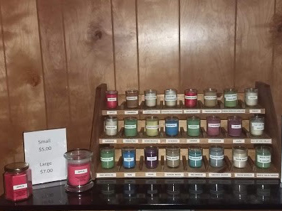 There is a wooden stand with three rows of 8 scented soy candles showing the selection of scents. (The labels are not readable but there are many colors.)