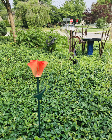 A red tulip shape sits upon a long stalk. In the background are other metal sculptures in a sea of green groundcover.