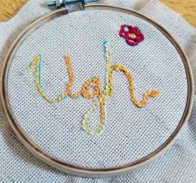 An embroidery ring, with an off-white piece of cloth stretched in it, has an embroidery of the word "Ugh!"