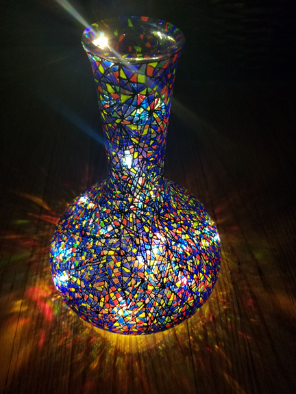 A glass vase with a round ball shape below and fluted tall above is mostly blue but with red and amber in a cut-glass stained-glass look. Fairy lights have been placed inside so it shines from within.