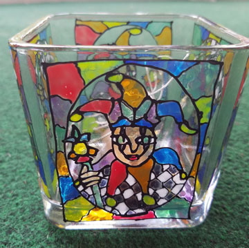 A tealight holder has been decorated to look like stained glass. It is a colorful joker, as if from a playing card or kid's book.